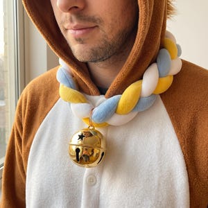May include: A braided plush collar featuring soft, padded fabric strands in yellow, light blue, and white. A large, polished gold-tone metal jingle bell with star and vertical slit cutouts hangs from the center of the braided fabric collar.