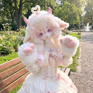 May include: A doll with white faux fur, pink horns, and a heart-shaped antenna on its head. It has large purple eyes, pink paw-shaped gloves, and wears a cream-colored dress with lace trim, pink ribbons, and a small cross pendant on the bodice.