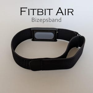 Op de afbeelding: Een zwarte stoffen armband met een rechthoekige zwarte behuizing met een grijs ovaal inzetstuk in het midden. De tekst FITBIT AIR en Bizepsband staat boven de band gedrukt. De band bevat een zwarte sluiting en een verstelbaar lusmechanisme.