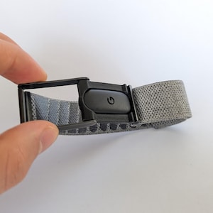 May include: A grey elastic belt featuring a black metal buckle mechanism. The buckle includes a central black rectangular component with a power symbol icon embossed on its surface. The belt strap has a textured, woven appearance in shades of grey.