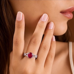May include: A silver-toned ring featuring a central oval-cut deep red ruby gemstone. The band is designed with a crisscross pattern and is encrusted with small, clear, sparkling stones along the metal surface, creating a detailed look.