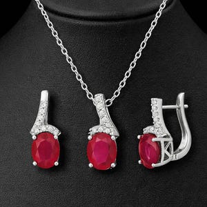 Könnte beinhalten: Ein Schmuckset bestehend aus einer silberfarbenen Halskette und passenden Ohrringen. Jedes Stück verfügt über einen großen, ovalen, tiefroten Edelstein, der mit einer Reihe kleiner, klarer, funkelnder Steine in einem silberfarbenen Rahmen besetzt ist.