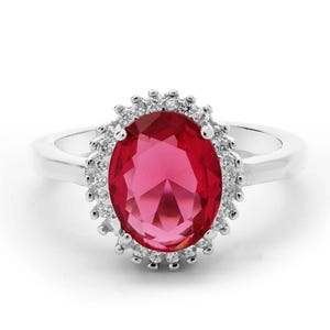 Puede incluir: Un anillo de plata con una gran piedra central de color rojo vibrante y corte ovalado. La gema central está rodeada por un halo de pequeñas piedras claras de corte redondo engastadas en un marco de plata festoneado sobre una banda lisa.
