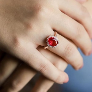 May include: A ring featuring a large, oval-cut, vibrant red gemstone set in a metallic band. The central red stone is surrounded by a halo of small, clear, sparkling stones, creating a classic and elegant design for this piece of jewellery.