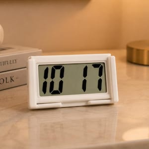 May include: A rectangular white digital clock with a clear LCD screen displaying the time 10:17 in bold black digits. The clock features a simple frame design with small protruding tabs at the bottom corners, resting on a smooth surface.