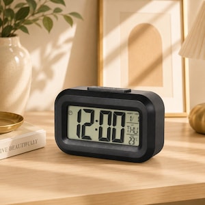 May include: A rectangular black digital alarm clock with a rounded frame. The screen displays the time 12:00 in large digits, along with the date 1/1, the day THU, and the temperature 23°C. A button is located on the top of the clock housing.