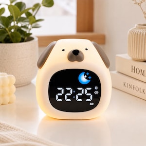 May include: A cream-coloured digital alarm clock shaped like a dog with brown ears and a dark brown nose. The front features a black screen displaying the time 22:25, a blue moon icon, a small alarm indicator, and the text Sat in white digital font.