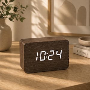 May include: A rectangular digital alarm clock with a dark brown wood grain finish. The front face displays the time 10:24 in bright white LED numerals. The clock has rounded corners and a smooth, solid construction, presenting a clean, functional design.