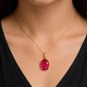 May include: A gold chain necklace featuring a single oval-cut vibrant red gemstone pendant. The pendant is secured in a simple four-prong setting, suspended from a delicate gold link chain that rests against the collarbone area.