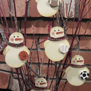 May include: Five wooden snowman ornaments with different sports-themed decorations. Each snowman is painted white with a red scarf and black eyes and mouth. The ornaments are hanging from a branch with a brown brick background.