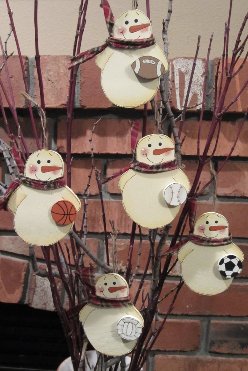 May include: Five wooden snowman ornaments with different sports-themed decorations. Each snowman is painted white with a red scarf and black eyes and mouth. The ornaments are hanging from a branch with a brown brick background.
