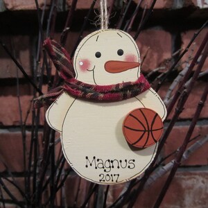 May include: A wooden snowman ornament with a red and black scarf, holding a basketball. The snowman has a painted face and the text "Magnus 2017" is written on the ornament.