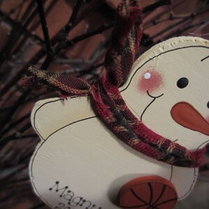 May include: A white snowman ornament with a red nose and black eyes. The snowman is wearing a red and black plaid scarf and has a basketball-themed decoration on its belly. The ornament is labeled "Magni" on the bottom.