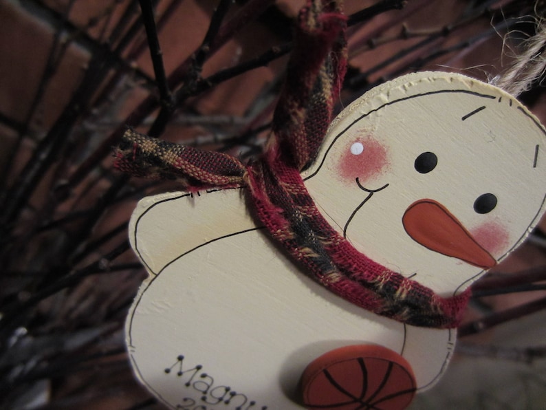 May include: A white snowman ornament with a red nose and black eyes. The snowman is wearing a red and black plaid scarf and has a basketball-themed decoration on its belly. The ornament is labeled "Magni" on the bottom.