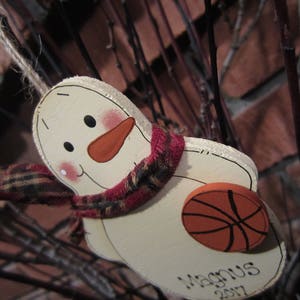 May include: A white wooden snowman ornament with a red, black, and white plaid scarf, a red nose, and a black basketball. The ornament is decorated with the name "Magnus" and the year "2017".