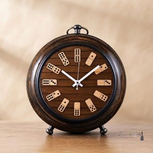 Op de afbeelding: Een ronde analoge klok met een donkerbruine houten rand en een wijzerplaat met horizontale planken. In plaats van traditionele cijfers heeft de klok twaalf lichtgekleurde houten dominostenen in een cirkel, met witte uur- en minutenwijzers.