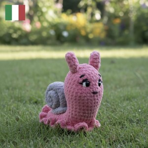 May include: A handmade crochet snail plush toy featuring a soft pink body with a textured, ruffled base and a light gray spiral shell. The snail has two upright antennae, black embroidered eyes with long lashes, and a small black stitched mouth.