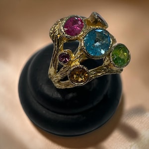 May include: A gold ring with a textured, branch-like band featuring multiple bezel-set faceted gemstones in various colours, including a large central blue stone, a pink stone, a green stone, a small purple stone, and a brown stone.