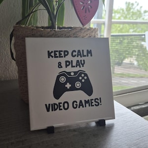 Può includere: Una piastrella quadrata in ceramica bianca con testo nero che recita KEEP CALM & PLAY VIDEO GAMES! centrato sopra e sotto un'illustrazione grafica nera di un controller per videogiochi. La piastrella è sostenuta da due piccoli supporti in plastica nera.