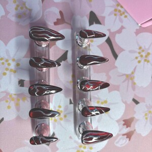 May include: A set of ten stiletto-shaped press-on nails featuring a deep red, glittery base with metallic silver vein-like patterns. The nails are arranged in two vertical rows on clear plastic stands, showcasing a sharp, pointed tip design.