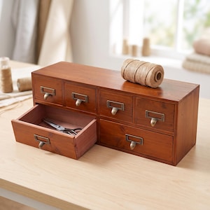 May include: A rectangular wooden desktop organizer with a medium brown finish, featuring five small drawers with antique brass-toned metal label pulls. One drawer is pulled open to reveal a pair of silver metal scissors inside the storage unit.