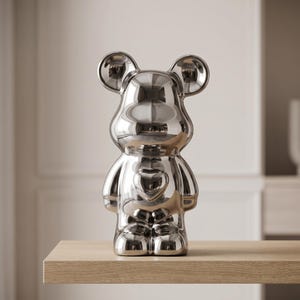 May include: A reflective silver-toned bear figurine with a smooth, mirror-like finish. The bear features rounded ears, a prominent heart-shaped indentation on its chest, and a stylized, anthropomorphic body shape with short arms and legs.