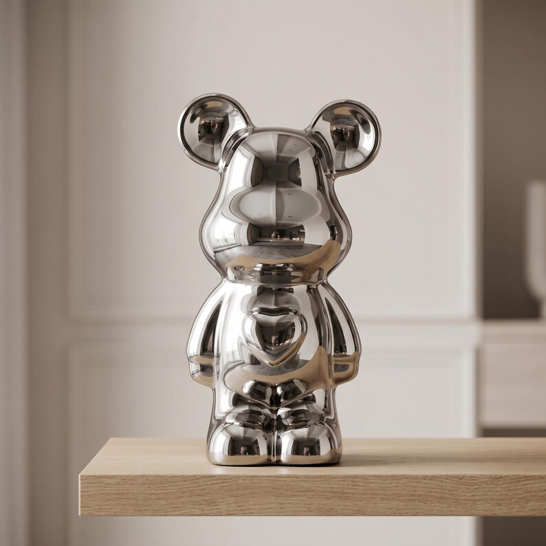 May include: A reflective silver-toned bear figurine with a smooth, mirror-like finish. The bear features rounded ears, a prominent heart-shaped indentation on its chest, and a stylized, anthropomorphic body shape with short arms and legs.