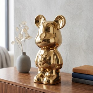 May include: A shiny gold-colored bear-shaped figurine with a smooth, reflective surface and a prominent heart-shaped detail on its chest. The bear has rounded ears, a rounded head, and a standing posture, crafted with a polished metallic finish.