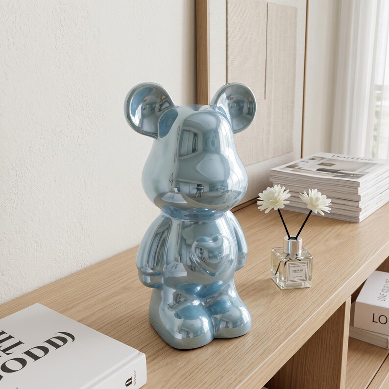 May include: A glossy, metallic light blue bear-shaped figurine with a smooth, reflective surface and a heart-shaped detail on its chest. The bear features rounded ears and a stylized, anthropomorphic form, standing upright as a decorative object.