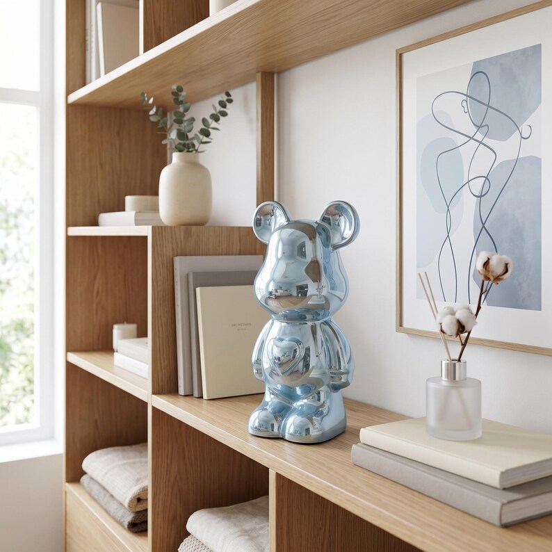 May include: A glossy, metallic light blue bear-shaped figurine with a smooth, reflective surface. The bear features rounded ears, a defined snout, and a heart-shaped detail on its chest, standing upright as a decorative object on a wooden shelf.