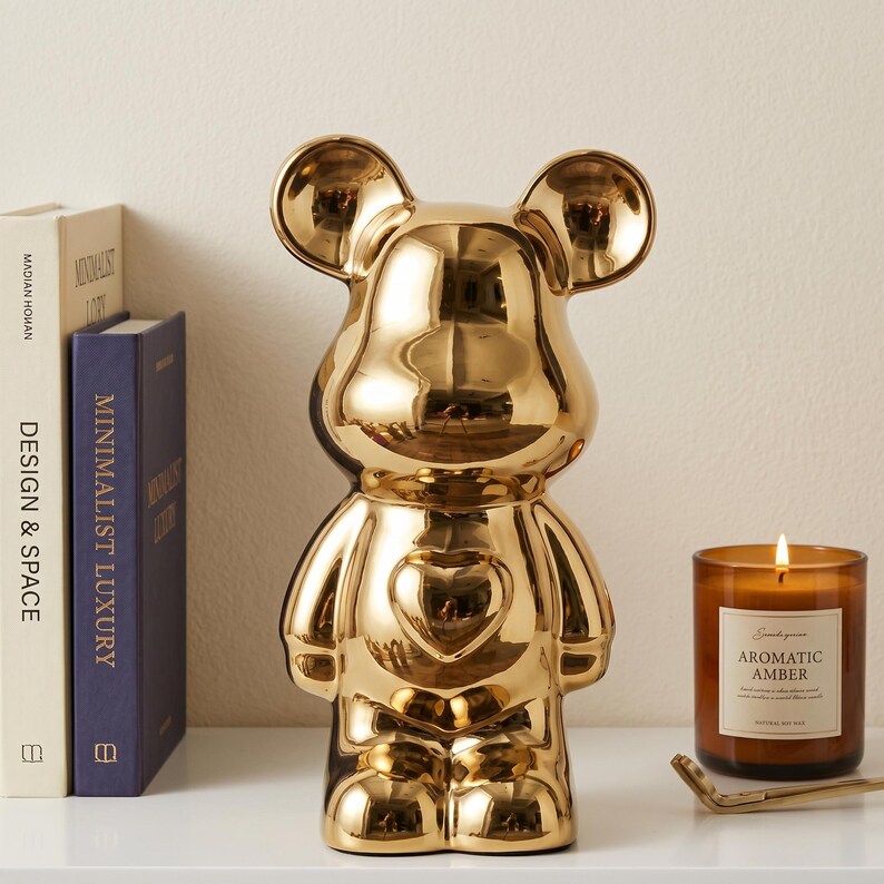 May include: A shiny gold-toned bear-shaped figurine with a heart-shaped detail on its chest. The bear has rounded ears and a smooth, reflective surface. It stands upright with a symmetrical design, featuring a stylized head and body.