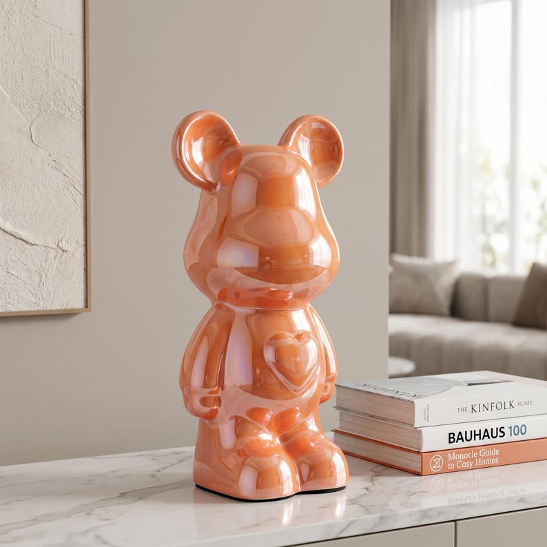 May include: A glossy, iridescent orange bear-shaped figurine with a smooth, reflective surface and a prominent heart-shaped detail on its chest. The bear features rounded ears and a stylized, anthropomorphic form standing in an upright position.