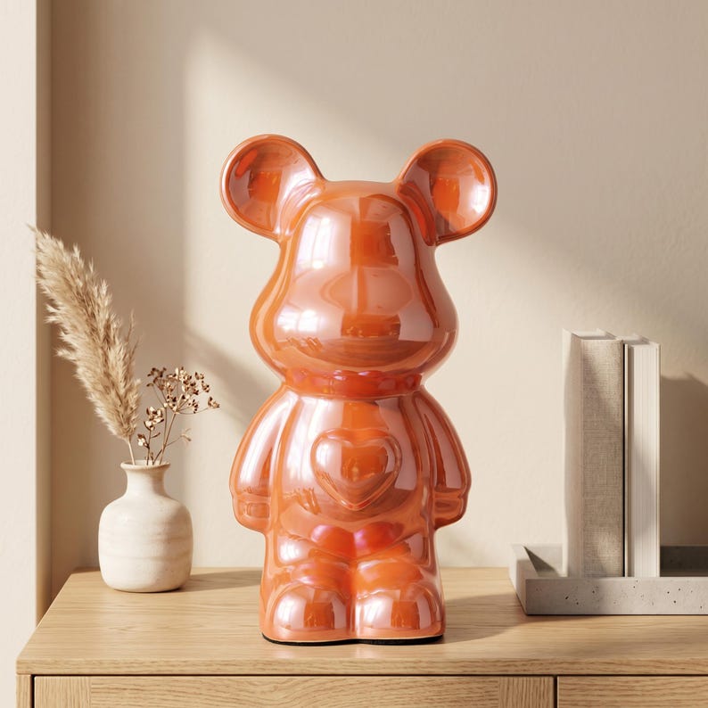 May include: A glossy, metallic orange bear-shaped figurine featuring a smooth, rounded silhouette with large ears and a prominent heart-shaped emblem centered on its chest. The surface has a reflective, iridescent finish that catches light.