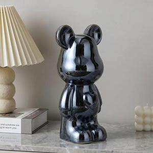 May include: A standing bear-shaped figurine with a smooth, reflective dark metallic finish. The bear features rounded ears, a smooth head, and a heart-shaped detail on its chest. The surface is highly polished, creating a mirror-like effect.