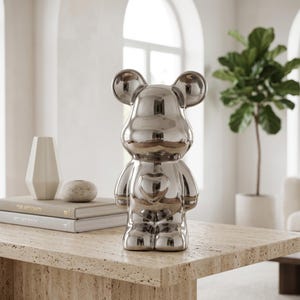 May include: A reflective silver-colored bear-shaped figurine with a smooth, metallic finish. The bear features rounded ears, a distinct snout area, and a prominent heart-shaped indentation centered on its chest, standing upright on a flat surface.