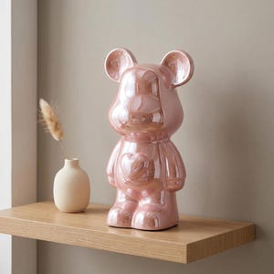 May include: A glossy, metallic pink bear-shaped figurine featuring a prominent heart embossed on its chest. The bear has rounded ears and a smooth, reflective surface. Beside it sits a small, matte beige vase containing a single dried grass stem.
