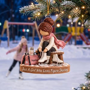 Op de afbeelding: Een hangend ornament met een figuur met bruin haar in een knot, gekleed in een crèmekleurige trui en roze sjaal, zittend op een houten bankje terwijl witte schaatsen worden gestrikt. Een roze tas met het label Sophia en een flesje staan ernaast. De tekst luidt: Just A Girl Who Loves Figure Skating.