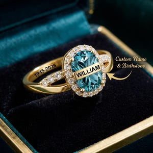 May include: A gold ring featuring an oval blue gemstone with the name WILLIAM engraved across the center. The band is accented with small clear stones and the text 1945-2026 is engraved on the side. The text Custom Name & Birthstone is visible.