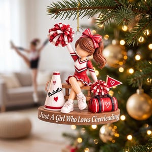 May include: A cheerleader ornament featuring a figure in a red and white uniform sitting on a wooden bench. The base reads "Just A Girl Who Loves Cheerleading" and includes a megaphone labelled "Isabella," a red duffel bag, and a pennant.