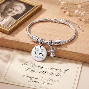 Könnte beinhalten: Ein silberfarbenes Schlangenkettenarmband mit einem runden Anhänger, der mit Engelsflügeln, dem Namen Tracy und den Daten 1955-2026 graviert ist. Das Armband enthält einen kleinen Engel-Anhänger und zylindrische Perlen mit dem Wort LOVE und einem Herz.