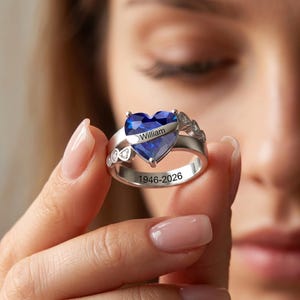 May include: A silver-toned ring featuring a large heart-shaped blue gemstone. A metal band across the center of the stone is engraved with the name William. The inner band is engraved with the dates 1946-2026. Small clear stones accent the sides.