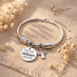 Op de afbeelding: Een zilverkleurige slangenkettingarmband met een ronde hanger gegraveerd met de naam William en de data 1955-2026, bekroond met engelvleugels. De armband bevat een kleine engelbedel en cilindrische kralen met het woord LOVE.