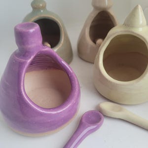 May include: Set of four ceramic salt cellars in purple, green, beige, and yellow with a small spoon. The salt cellars are shaped like small houses with a rounded top and a small opening for pouring salt.