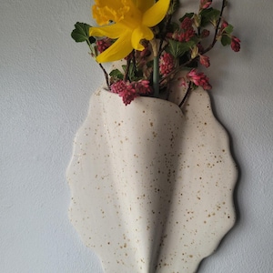 May include: White ceramic wall vase with a speckled glaze and a unique, wavy shape. A yellow daffodil and pink flowers are arranged in the vase.