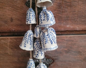 May include: A string of ceramic bells with a blue and white floral design. The bells are suspended from a natural fiber rope. The bells are of varying sizes and shapes, with a glossy finish.