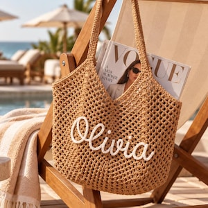 May include: A tan crochet tote bag featuring an open-weave design and two shoulder straps. The front of the bag displays the name Olivia in white cursive script lettering. A magazine is tucked inside the bag, partially visible at the top.