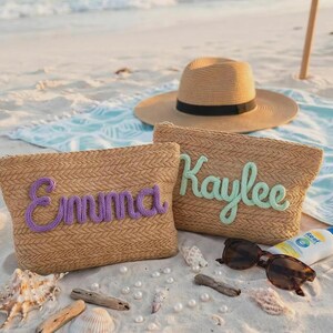 Puede incluir: Dos bolsos rectangulares de paja con textura tejida. Uno lleva el nombre Emma en letras de hilo morado, mientras que el otro muestra el nombre Kaylee en letras de hilo verde claro. Se incluye un sombrero de paja de ala ancha con una banda negra.