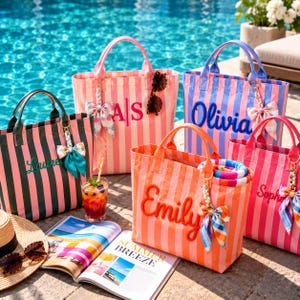 Puede incluir: Cuatro bolsos tote rectangulares con rayas verticales en rosa, verde, naranja y azul. Cada bolso presenta un nombre o iniciales personalizados en relieve, un llavero de lazo decorativo y dos asas. Una bebida y una revista están cerca.