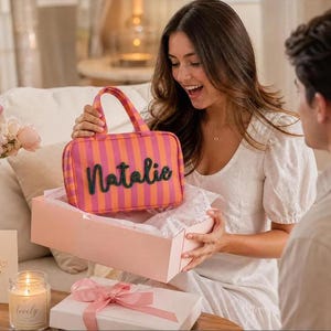 May include: A rectangular zippered pouch with a pink and orange vertical striped pattern and two top handles. The name Natalie is written in dark green, raised, cursive lettering across the front of the pouch, which is held above a light pink gift box.