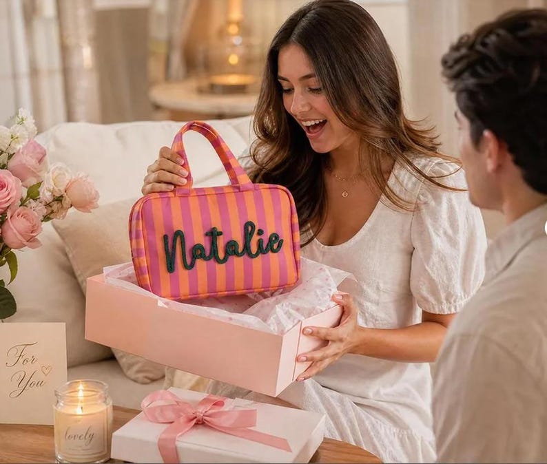May include: A rectangular zippered pouch with a pink and orange vertical striped pattern and two top handles. The name Natalie is written in dark green, raised, cursive lettering across the front of the pouch, which is held above a light pink gift box.